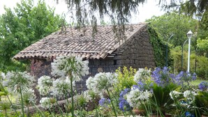 A stone building with a tiled roof surrounded by lush greenery and various flowering plants.