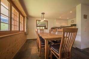 A dining area with a wooden table and chairs, a kitchen in the background, and a view of the outdoors through large windows.