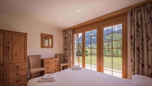 A bedroom with a large bed, wooden wardrobe, a mirror, and a view of a green landscape through large windows.