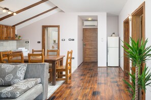 A compact kitchen with wooden cabinets, a dining table, and chairs. There is a sofa with patterned cushions, a potted plant, and a wooden-framed door.