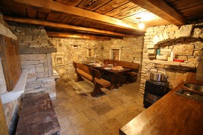 A rustic stone-walled kitchen with a wooden dining table, benches, and a built-in stove.