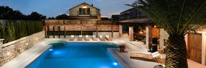 A swimming pool with lounge chairs, a stone wall, and a building with a tiled roof in the background.