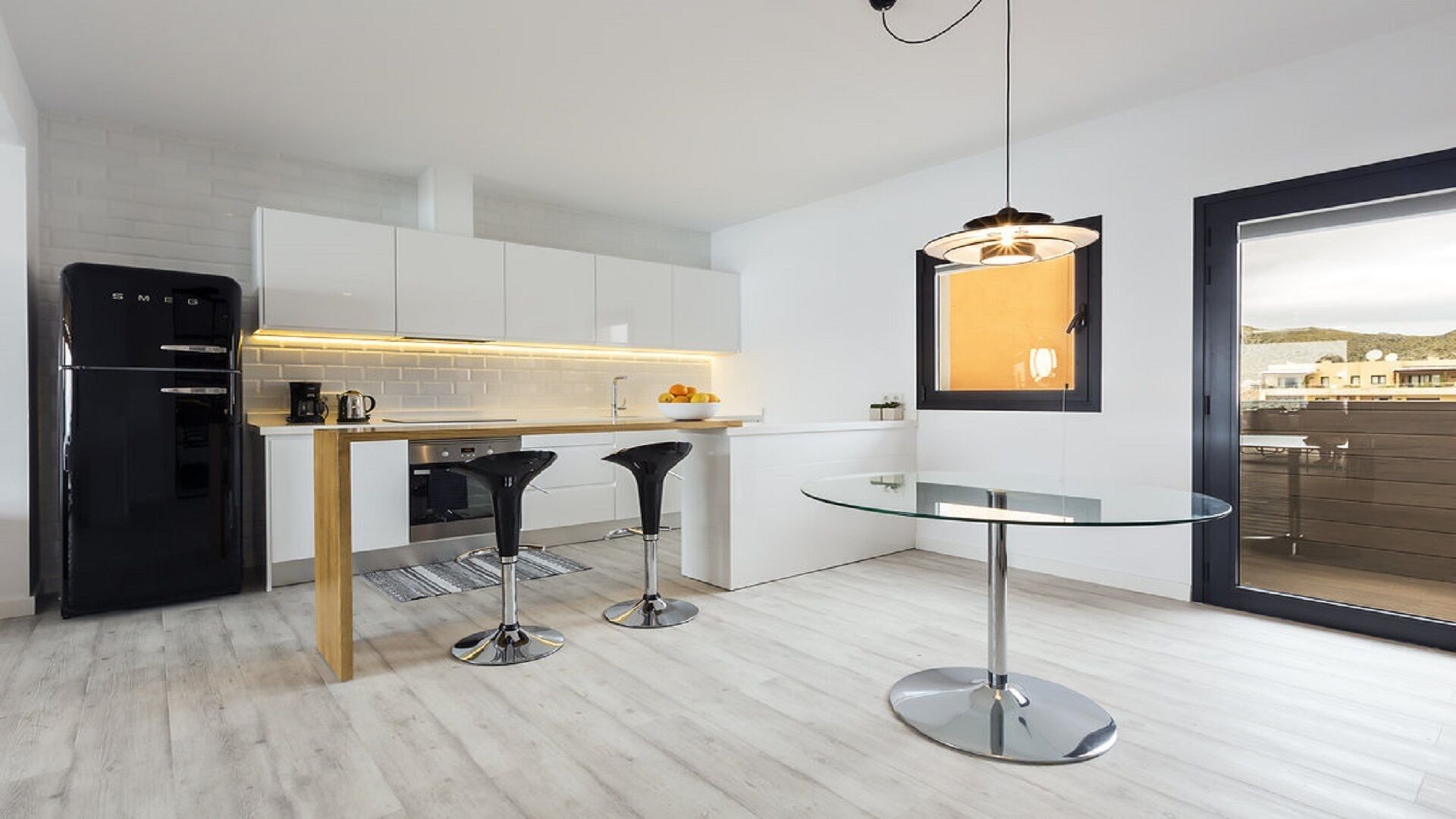 Een moderne keuken met witte kasten, een zwarte koelkast, een glazen eettafel en twee zwarte barkrukken.