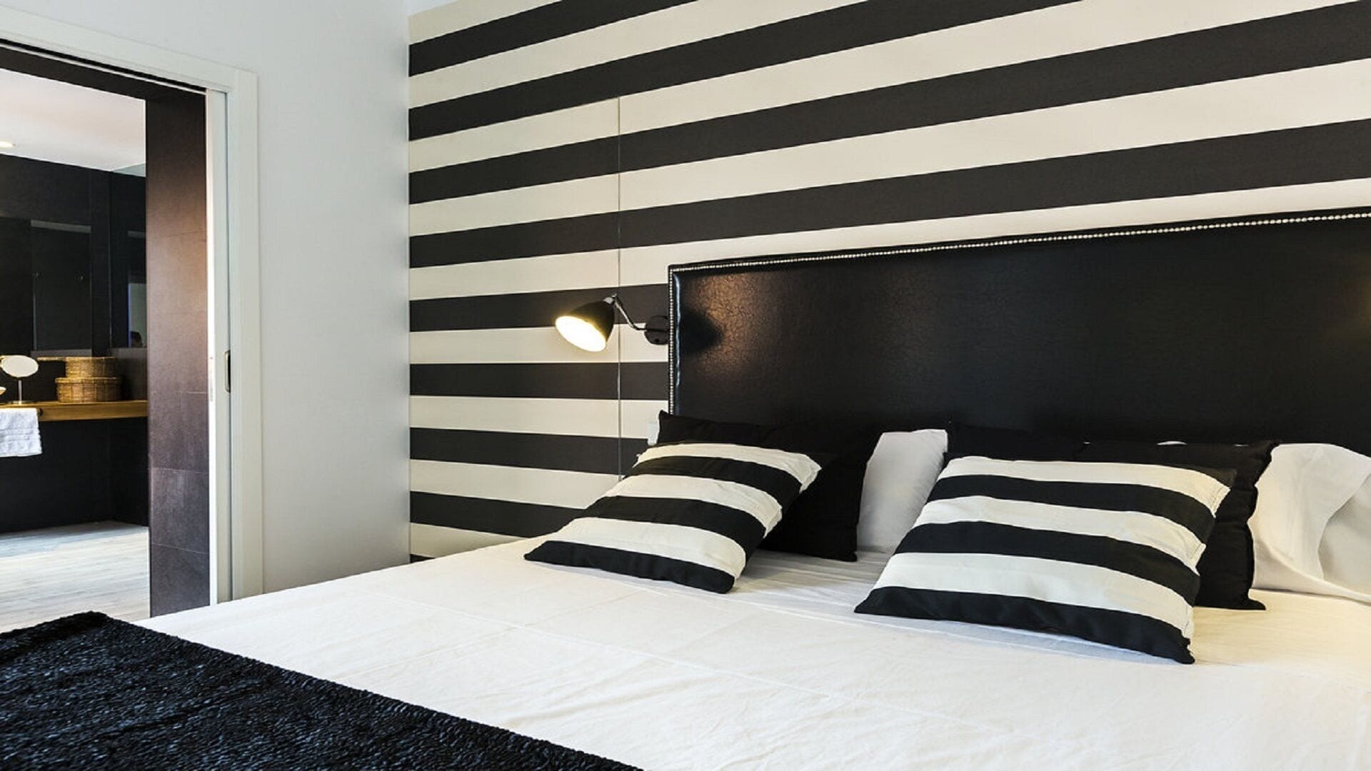Une chambre moderne avec un mur à rayures noires et blanches, un grand lit avec des oreillers à rayures noires et blanches, et une lampe de chevet.