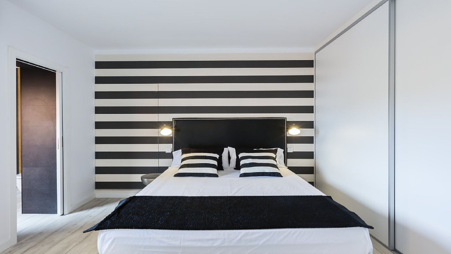 A modern bedroom with a bed centered against a striped wall, two bedside lamps, and a door to the left.