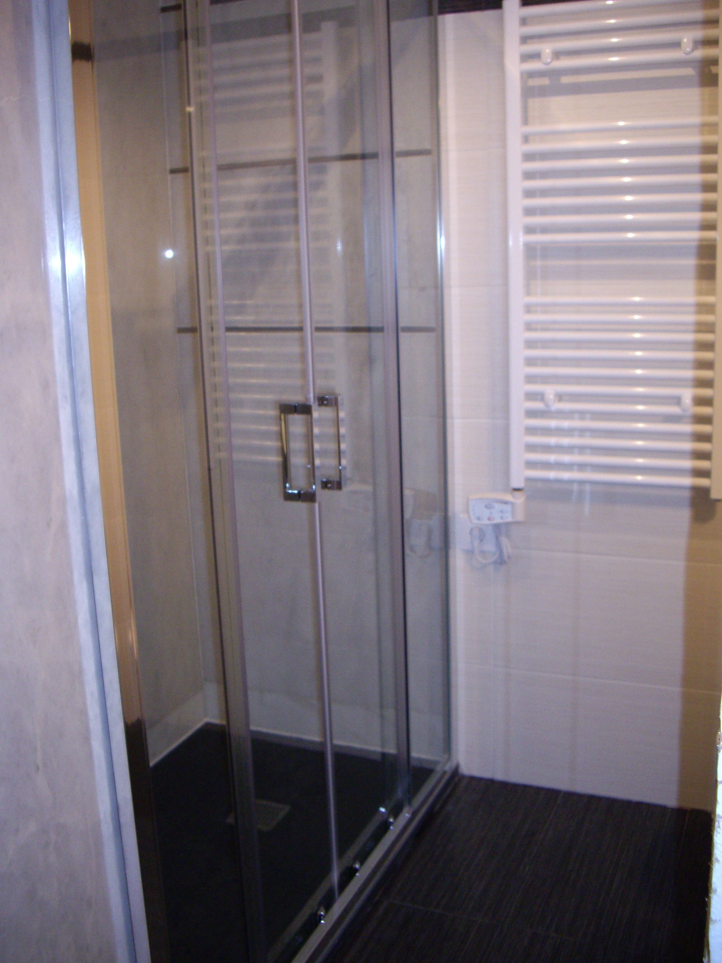 Una ducha con cerramiento de vidrio, un panel de control, pared con azulejos blancos y una ventana con persianas.