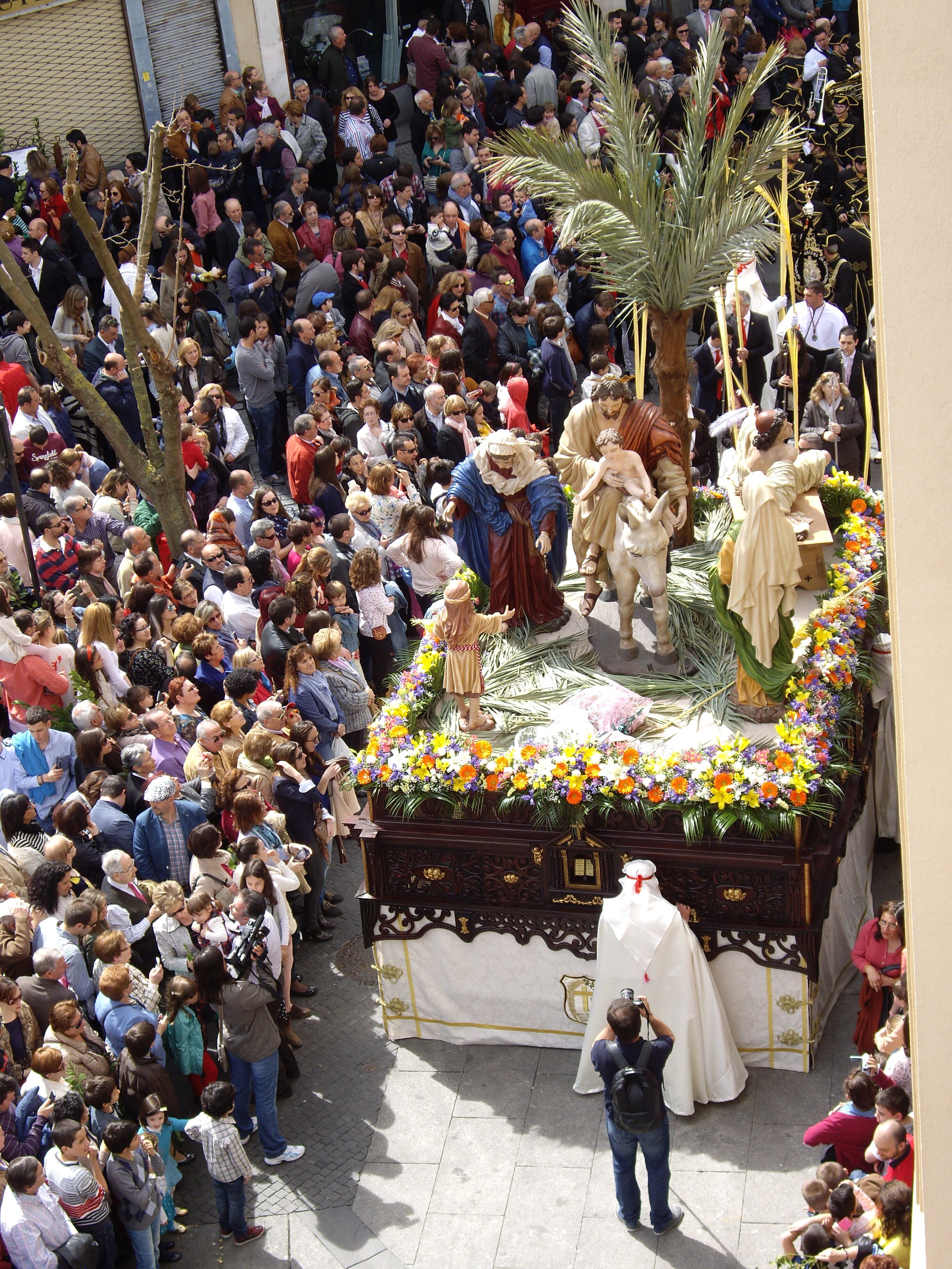 A crowded outdoor event with a central display featuring statues and floral arrangements.