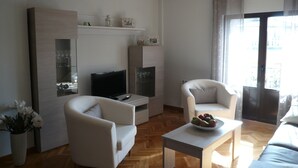 Ein modernes Wohnzimmer mit einem Flachbildfernseher, zwei weißen Sesseln, einem Couchtisch mit Obst und einem Holzboden.