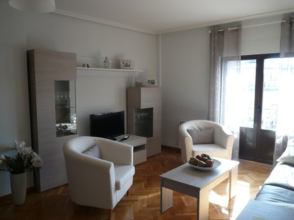 Un salon moderne avec un téléviseur à écran plat, deux fauteuils blancs, une table basse avec des fruits et un plancher en bois.