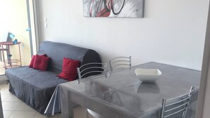A modern living room with a grey sofa, red cushions, a glass dining table with metal chairs, and a large abstract painting on the wall.
