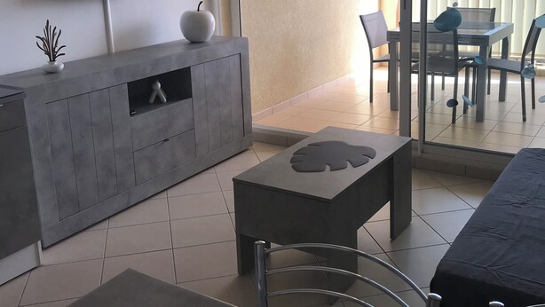 A modern living room with a flat-screen TV, a small white vase, and a decorative apple on a grey console table. There is a dining area with a table and chairs, and a balcony with a glass door leading outside.