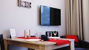 A hotel room with a dining table set for two, a flat-screen TV mounted on the wall, and a "HOME" sign above the TV.