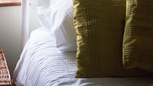 A bed with white and green pillows, a window with a view of greenery, and a wicker chair.