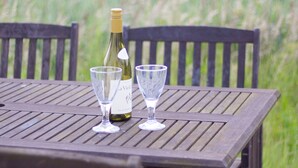 A wooden table with two wine glasses and a bottle of wine, set outdoors with a grassy field in the background.