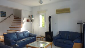 A living room with two blue leather sofas, a glass coffee table, a wooden side table, a black wood-burning stove, a ceiling fan, a window with a view of greenery, a staircase, and a bookshelf with books. - Lovely, two bedroom, air-conditioned Town House, set on a wonderful development (Paphos)