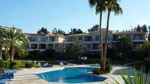 A pool area with lounge chairs, palm trees, and a building with balconies in the background. - Lovely, two bedroom, air-conditioned Town House, set on a wonderful development (Paphos)