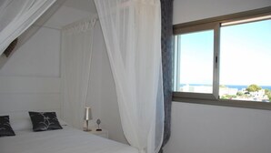 A bedroom with a bed, white curtains, a bedside table with a lamp, and a window with a view of buildings and the sea.