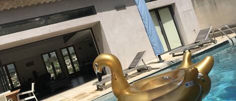 Uma piscina com dois brinquedos flutuantes de cisne dourado, cadeiras de espreguiçadeira e um edifício com telhado cerâmico ao fundo.