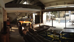 A spacious living area with a large sofa, a coffee table, and a dining table. There is a kitchen area in the background, and large windows provide a view of the outdoors. - At the top of the village, magnificent view of Mt Ventoux, private heated swimming pool (Bédoin)