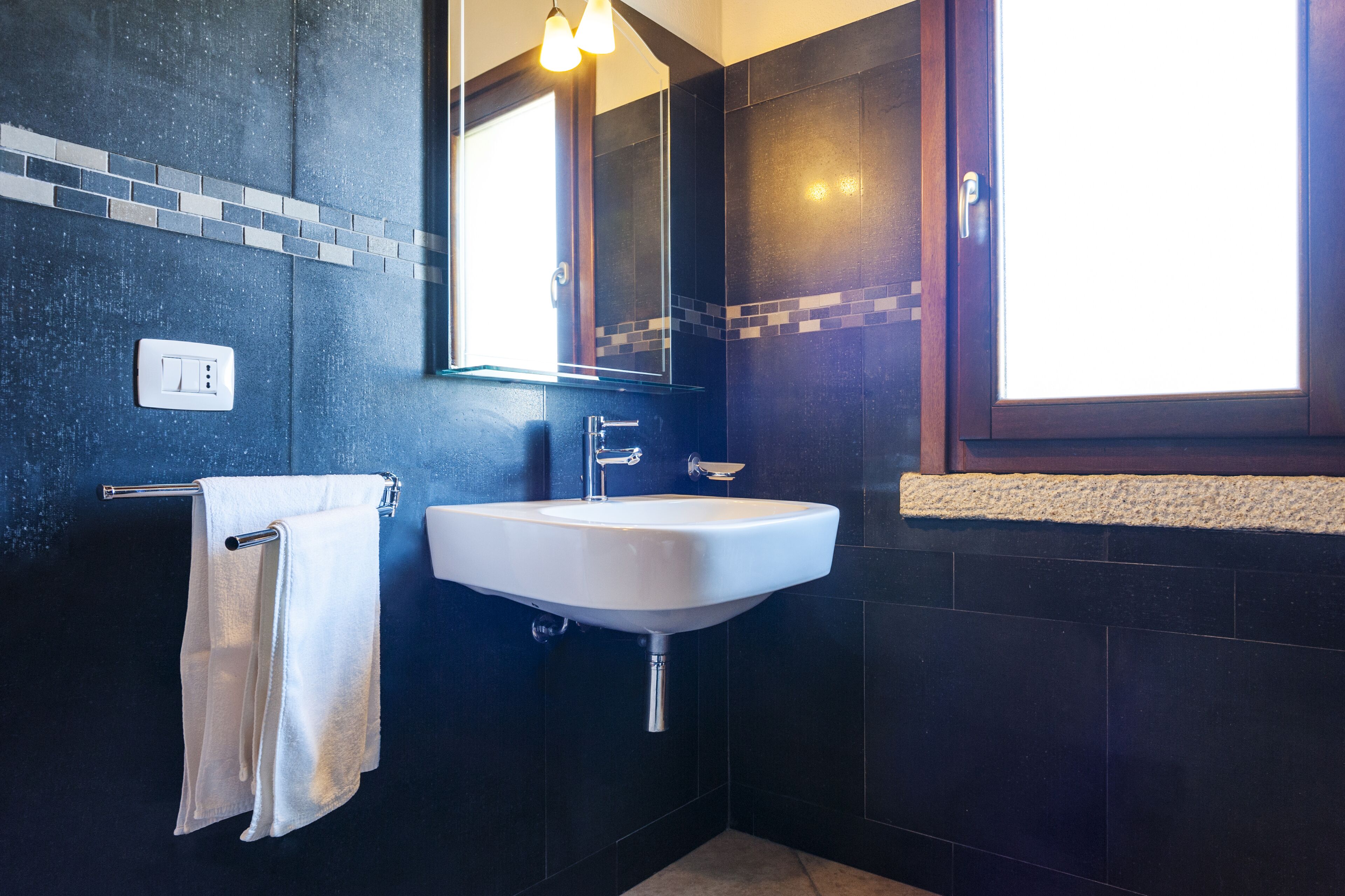 Een moderne badkamer met een wit wastafelmeubel, donkere betegelde wanden, een spiegel en een handdoekrek.