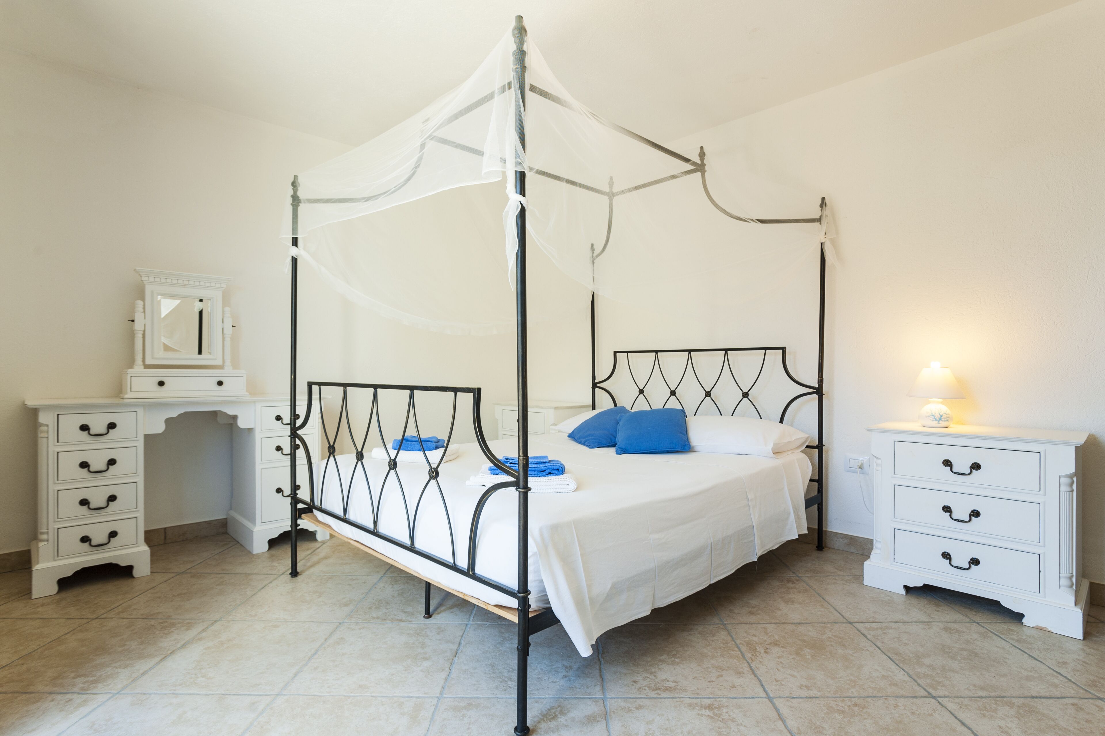 Una cama con dosel y cortinas, una mesita de noche con lámpara, y un aparador con cajones.