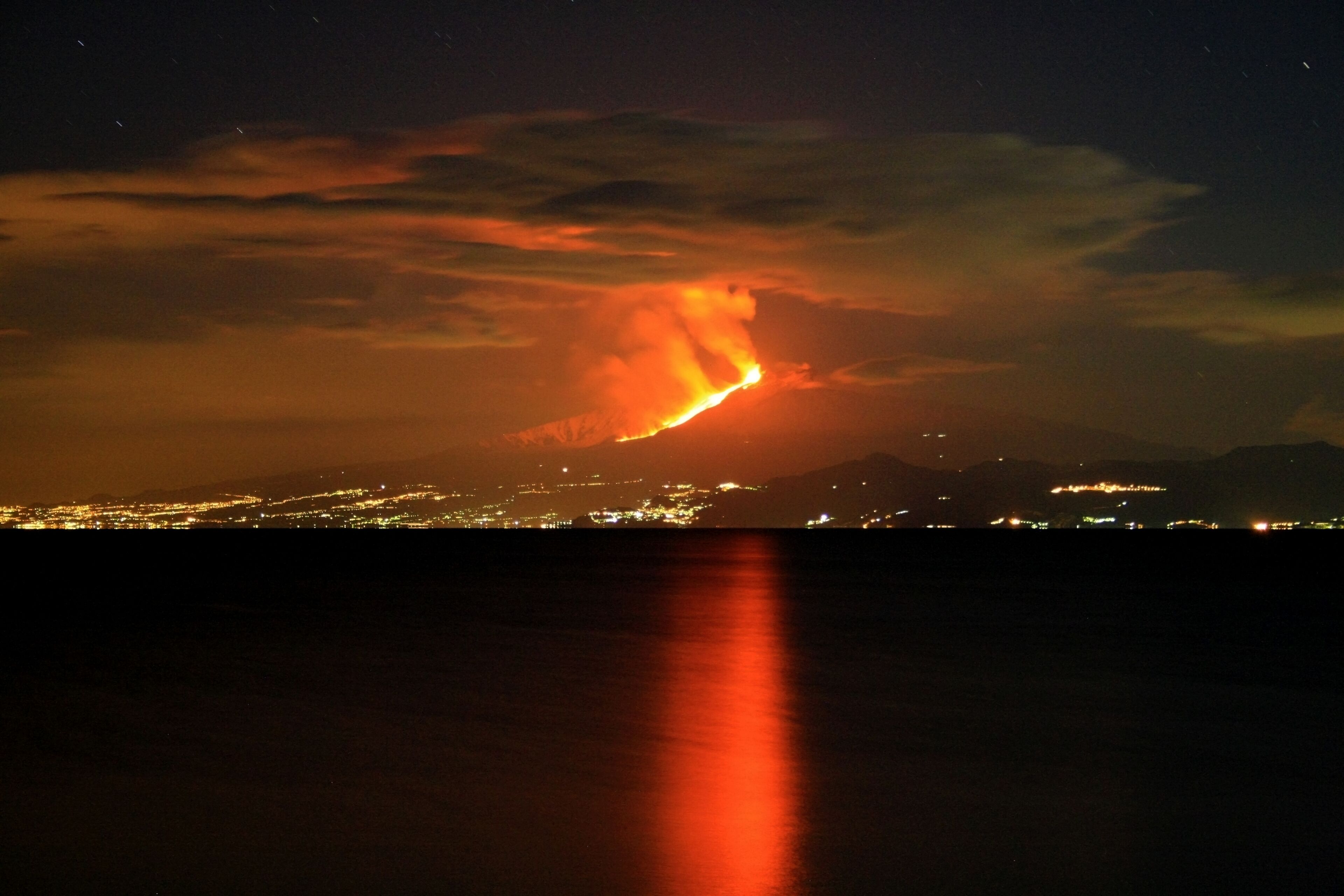 Une vue nocturne d’une ville, avec un volcan lointain en éruption et crachant de la lave.