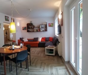 Un salon comprenant un canapé, une table basse et une table à manger avec des chaises. Un téléviseur est fixé au mur et une fenêtre laisse entrer la lumière naturelle.