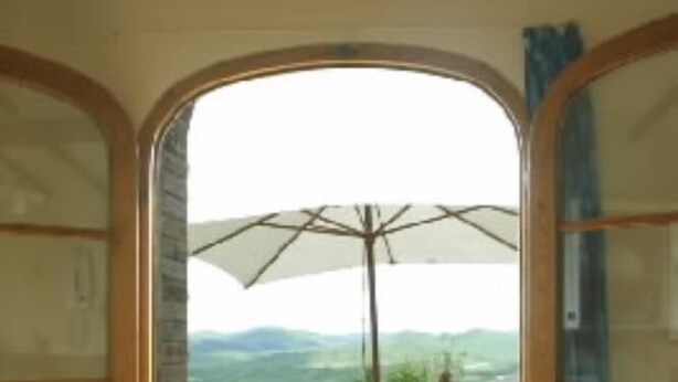 Un acceso abierto que da a un patio al aire libre con una mesa, sillas, un gran paraguas y vistas a un paisaje verde.