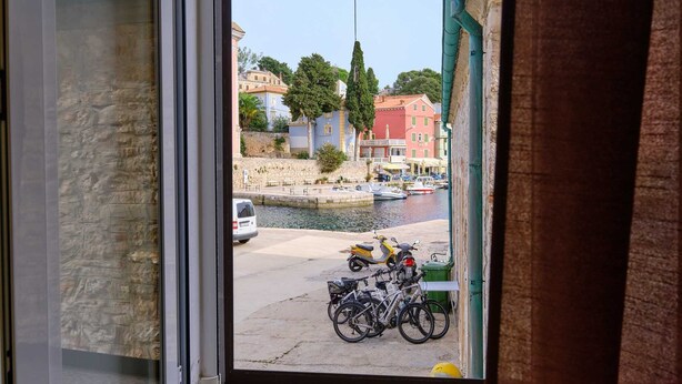 Une vue à travers une fenêtre donnant sur un port, avec des bateaux, des vélos et un mur de pierre.