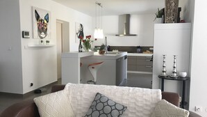 Un salon moderne avec un canapé marron, un fauteuil blanc et une table basse. On y trouve également un coin cuisine doté d’un plan de travail blanc et d’un four encastré.