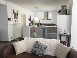 Un salon moderne avec un canapé marron, un fauteuil blanc et une table basse. On y trouve également un coin cuisine doté d’un plan de travail blanc et d’un four encastré.