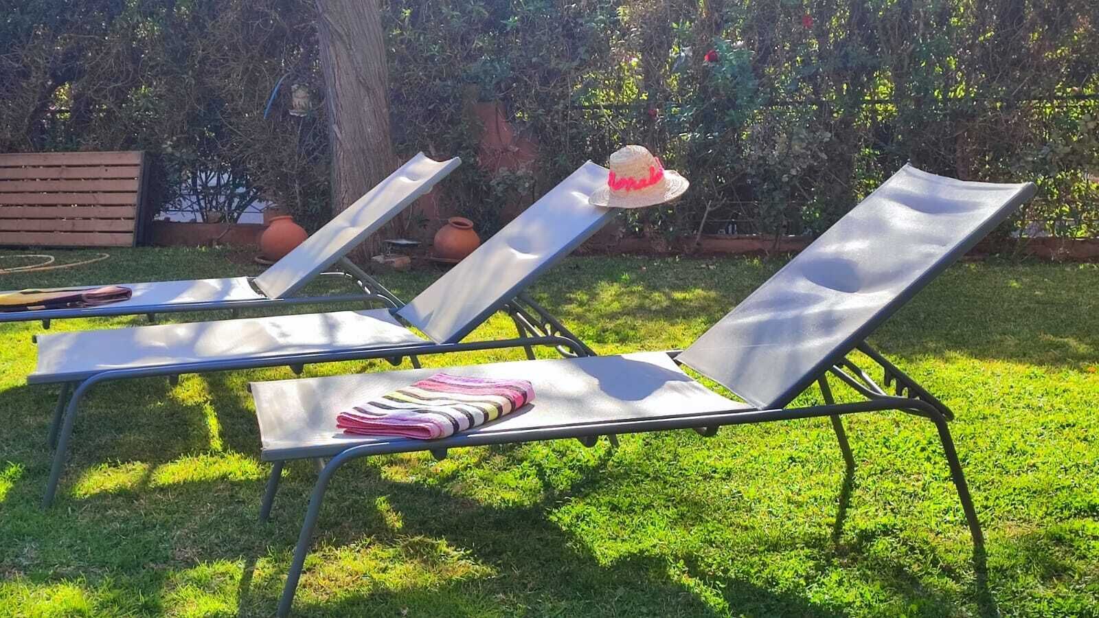 Trois chaises longues en métal avec des coussins, un chapeau et une serviette à rayures sur l’herbe.