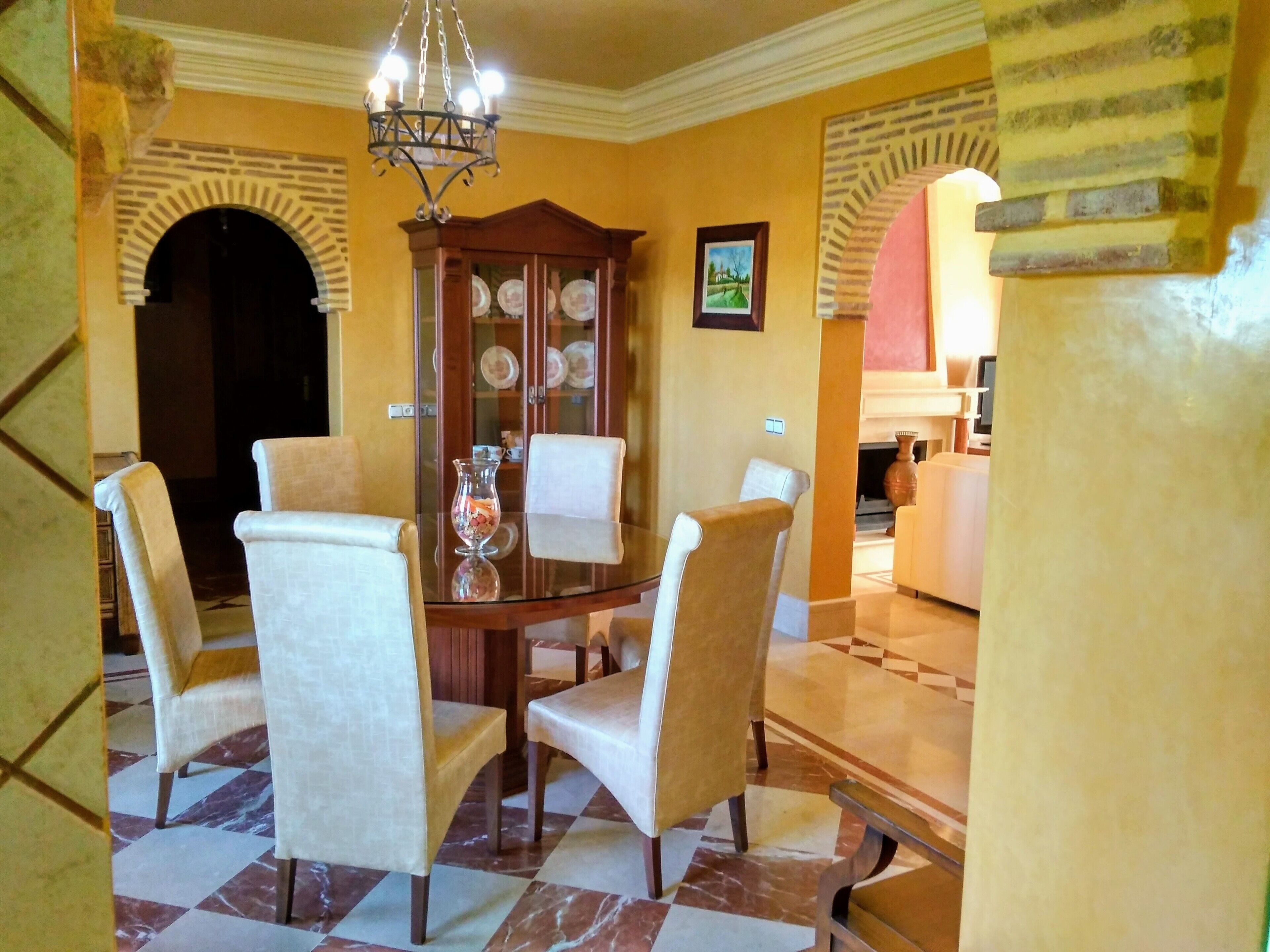 Un coin repas comprenant une table ronde et six chaises, une vitrine contenant de l’argenterie et un lustre.
