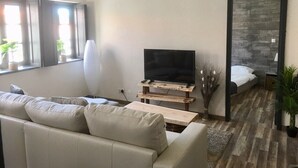 Un salon moderne avec un canapé, un téléviseur posé sur un meuble en bois, une petite table et un lit doté d’une tête de lit en bois.