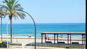 Blick auf einen Strand mit einer überdachten Sitzgelegenheit, Palmen und einem strahlend blauen Himmel.