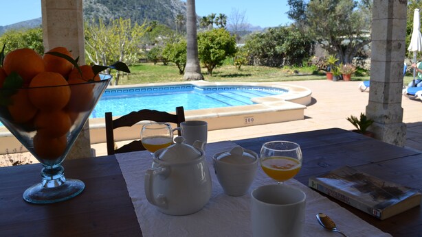 A table with a glass bowl of oranges, a white teapot, cups, and a book, overlooking a pool and mountains. - Villa ideal for families with pool and bbq near the sea in Pollença and WIFI (Pollença)