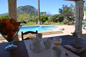 Une table sur laquelle se trouvent un saladier en verre rempli d’oranges, une théière blanche, des tasses et un livre, avec une vue sur une piscine et des montagnes.