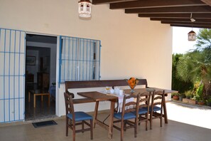 Une terrasse ouverte avec une table et des chaises en bois, une petite table avec une carafe et des verres, et une vue sur un jardin agrémenté de plantes en pot et d’un palmier.