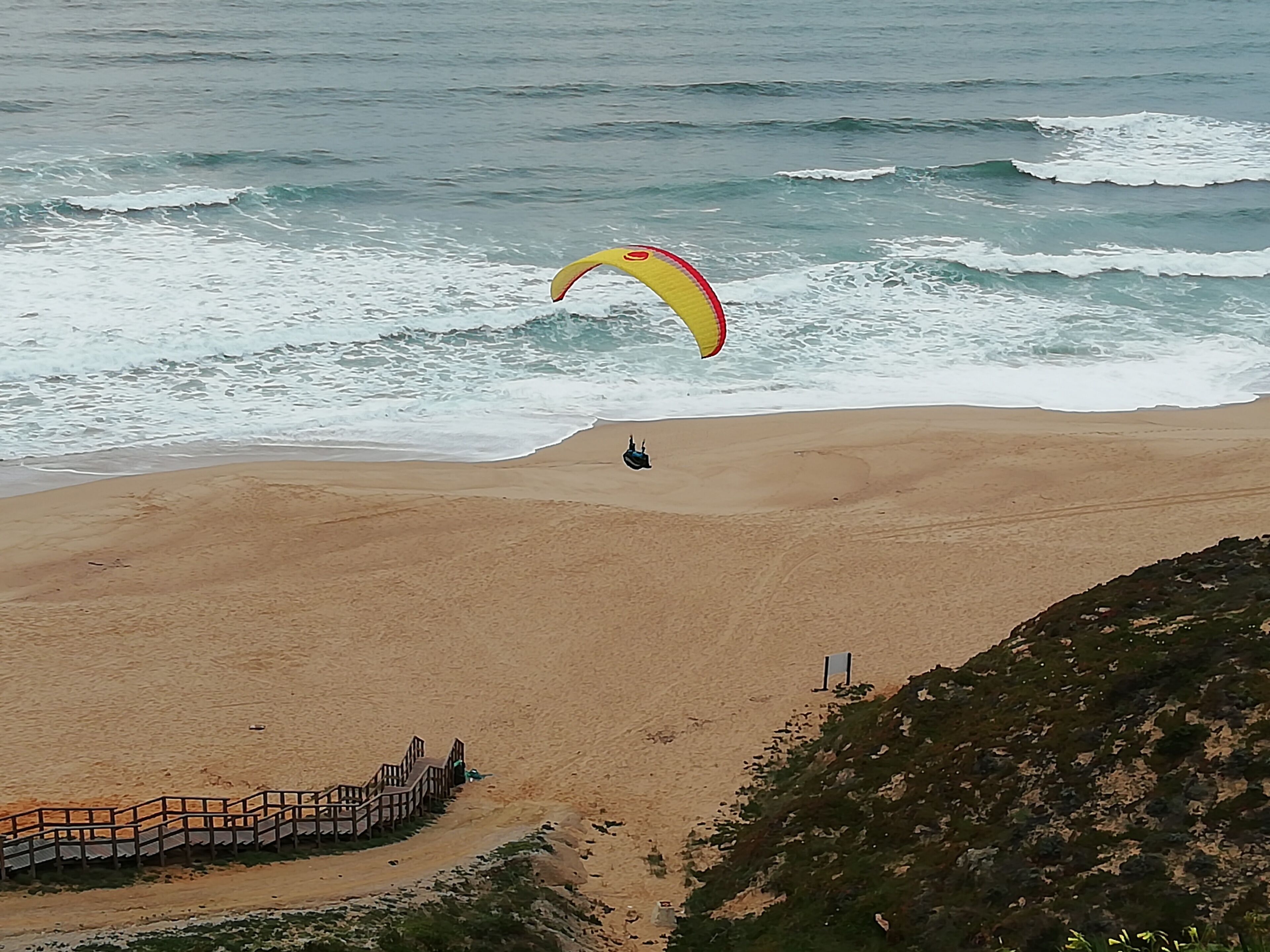 A beach with a wooden walkway leading to the water, a person parasailing, and a rocky outcrop in the foreground.