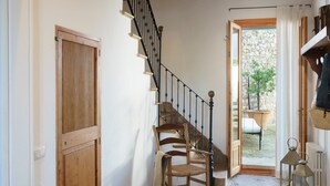 Une chaise en bois près d’un escalier avec une rampe noire, une porte en bois et une vue sur un jardin par une porte ouverte.