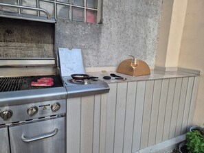 Un barbecue d’extérieur avec une plaque de cuisson et une spatule, un verre rempli de boisson et un panier en osier posé sur une plateforme en bois.