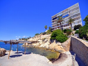 Hotel à beira-mar com costa rochosa, mar azul cristalino e edifício moderno com varandas viradas para o oceano.