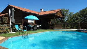 A swimming pool with a wooden deck, a covered patio area with chairs, and a wooden house in the background. - Timber frame house with garden (Vers)