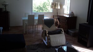 A room with a dining table and chairs, a rocking chair, a dresser, and a window with a view of trees. - Timber frame house with garden (Vers)