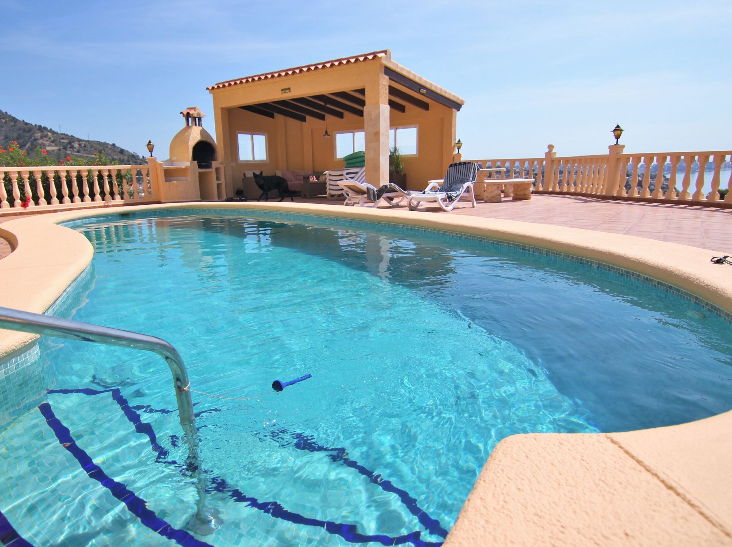 A swimming pool with a sunshade structure, lounge chairs, and a view of a hill.