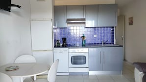 A compact kitchen with a white refrigerator, a stove, a sink, and a coffee maker. There is a small dining table with two chairs. - T2, 350 m from Bodri BEACH, Sea view, Air-conditioning, on large landscaped grounds. (Corbara)