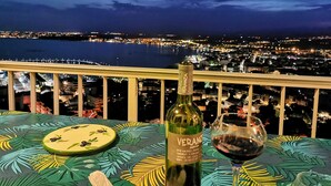 En flaska Verancia-vin och ett glas står på ett bord med ett tropiskt bladmotiv, med utsikt över en stadssiluett i nattljus.