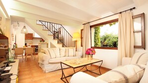 A spacious living area with a sofa, a coffee table, and a dining table. A staircase leads to an upper level. - Private Pool & Garden, BBQ, AC, PS4 and Views of the Tramuntana Mountains (Pollença)