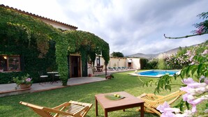 A backyard with a pool, wooden lounge chairs, a table, and a house with a tiled roof and greenery. - Private Pool & Garden, BBQ, AC, PS4 and Views of the Tramuntana Mountains (Pollença)
