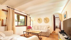 A living room with a sofa, a coffee table with flowers, a TV, and a view of the outdoors. - Private Pool & Garden, BBQ, AC, PS4 and Views of the Tramuntana Mountains (Pollença)